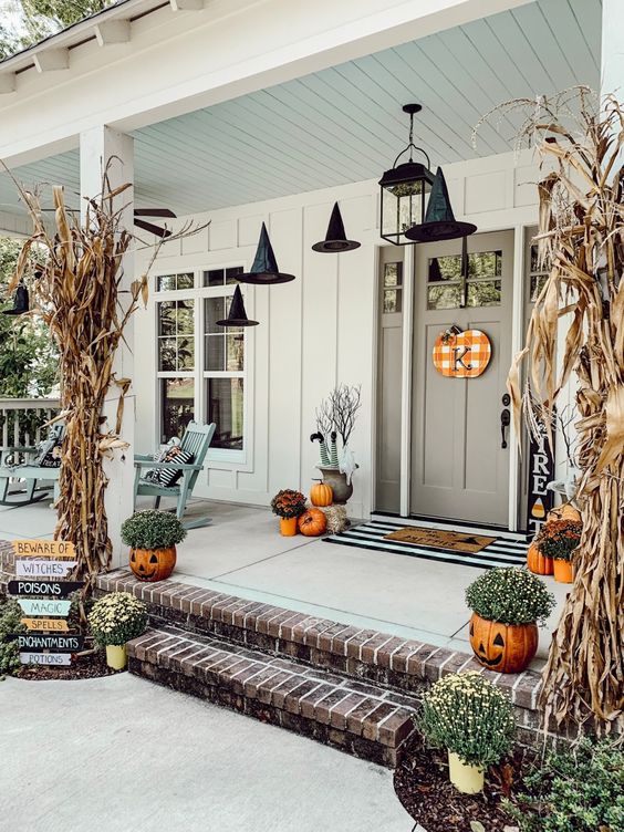 a cozy rustic Halloween porch with dried corn husks, pumpkins, pumpkin planters with blooms and witches' hats
