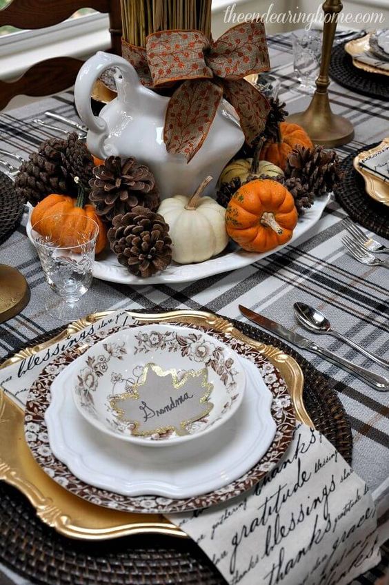 a cozy rustic vintage Thanksgiving tablescape with a plaid tablecloth, quote napkins, woven chargers, gold and pritned plates, a chic centerpiece of wheat, pumpkins and pinecones