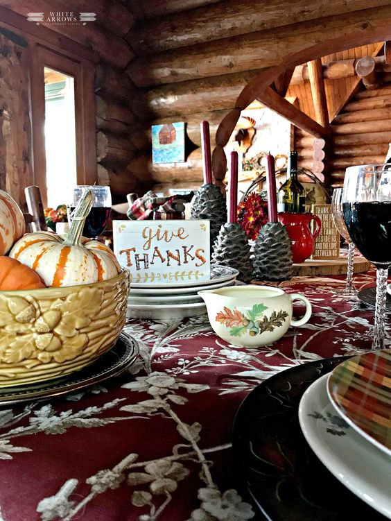 a cozy vintage-inspired Thanksgiving tablescape with a floral tablecloth, printed plates, a floral milk jug, pinecone candleholders and a bowl with pumpkins