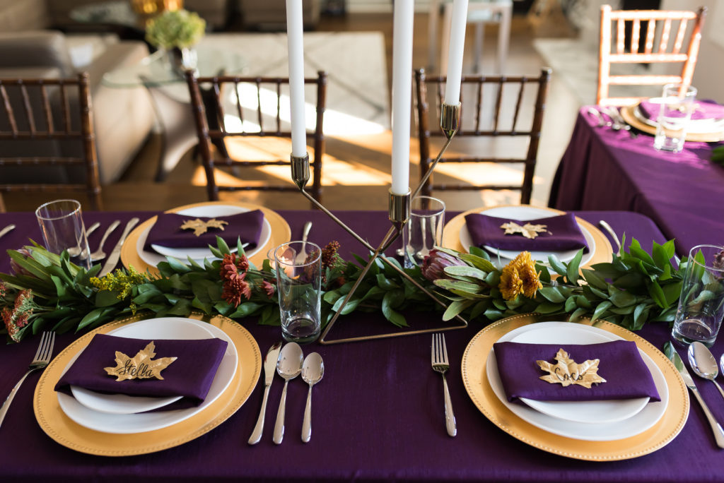 a gorgeous purple Thanksgiving tablescape with a purple tablecloth and napkins, a greenery and bright bloom runner, tall candles, gold touches and fall leaves