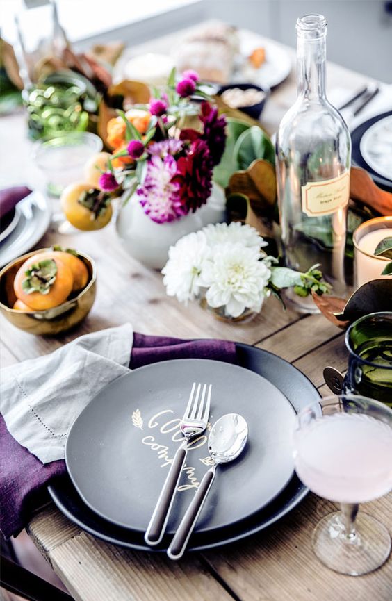 a laid-back and rustic Thanksgiving tablescape with greenery, bold purple blooms, fruits, black plates and purple napkins