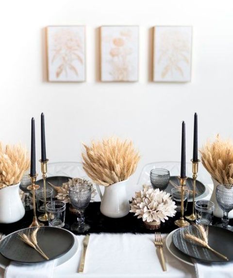 a modern glam Thanksgiving tablescape with wheat in vases, black candles and plates, a black runner and gold cutlery