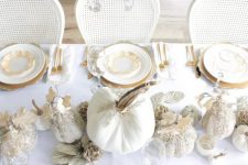 a refined white and gold Thanksgiving tablescape with white embellished pumpkins, white leaves, gold chargers and cutlery plus a crystal chandelier over the table