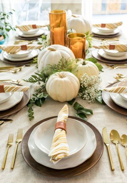 a rustic Thanksgiving table with wooden chargers, a burlap tablecloth, gold cutlery, white pumpkins and greenery plus candles