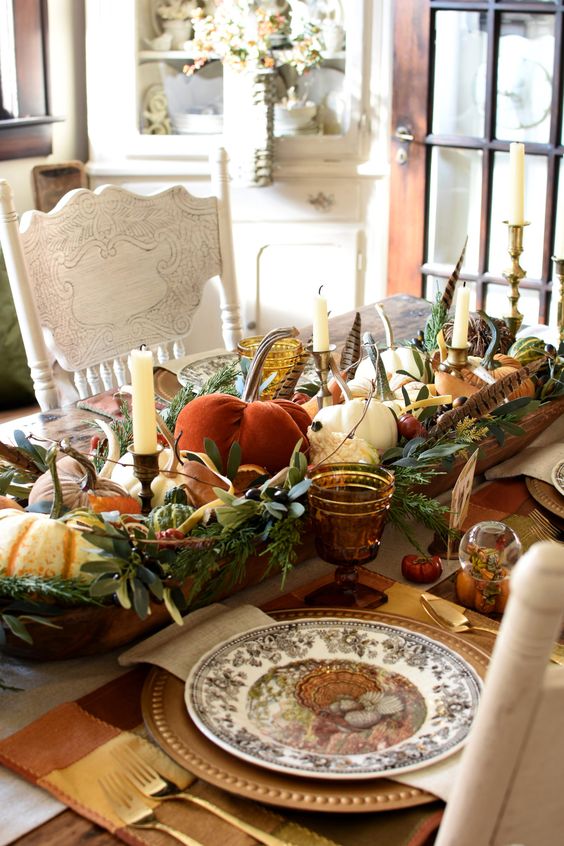 a rustic vintage Thanksgiving tablescape with a large bowl centerpiece with pumpkins, greenery, candles and feathers, plaid placemats, printed plates