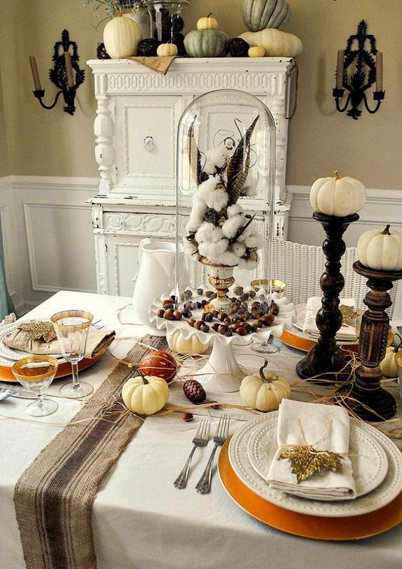 a rustic vintage Thanksgiving tablescape with neutral textiles, a striped runner, white porcelain and wooden stands with pumpkins plus cotton in a cloche