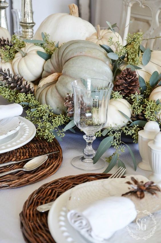 a rustic vintage Thanksgiving tablescape with woven chargers, white porcelain and textiles, natural pumpkins, greenery and pinecones