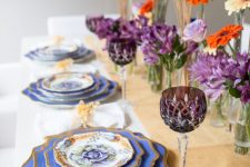 a stylish Thanksgiving table setting with a neutral runner, purple and orange blooms, wheat, blue plates with a gold edge, gold cutlery