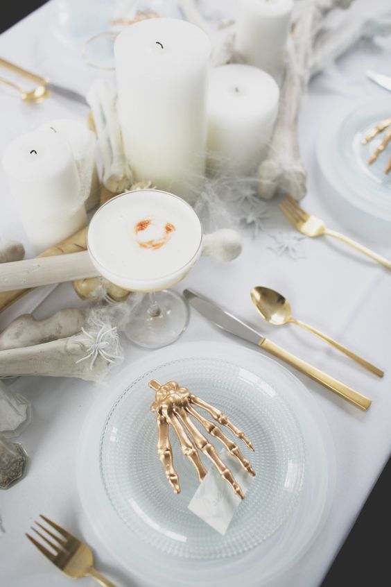 a white ghostly Halloween tablescape with bones, skeleton hands, pillar candles, gold cutlery and white drinks