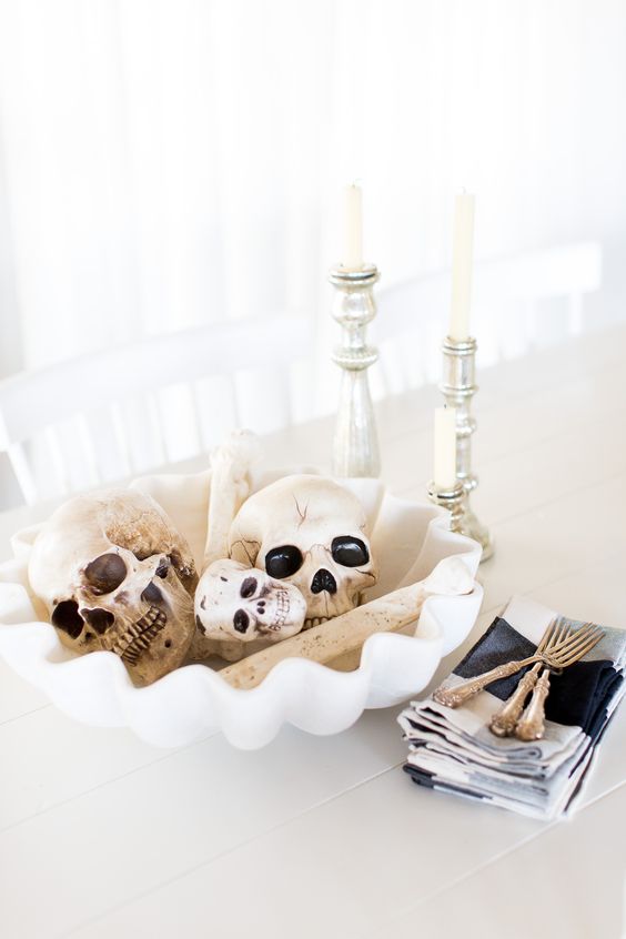 pretty white Halloween decor with a white bowl with skulls and bones, silver candleholders and white candles in them