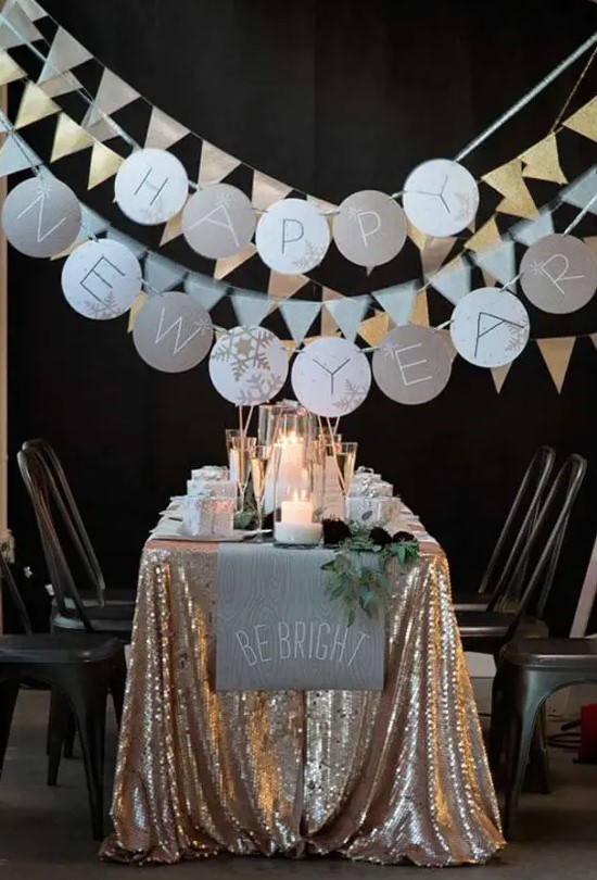a NYE party tablescape with a gold sequin tablecloth, a neutral table runner and greenery, candles and sparkling garlands is a gorgeous space to celebrate NYE