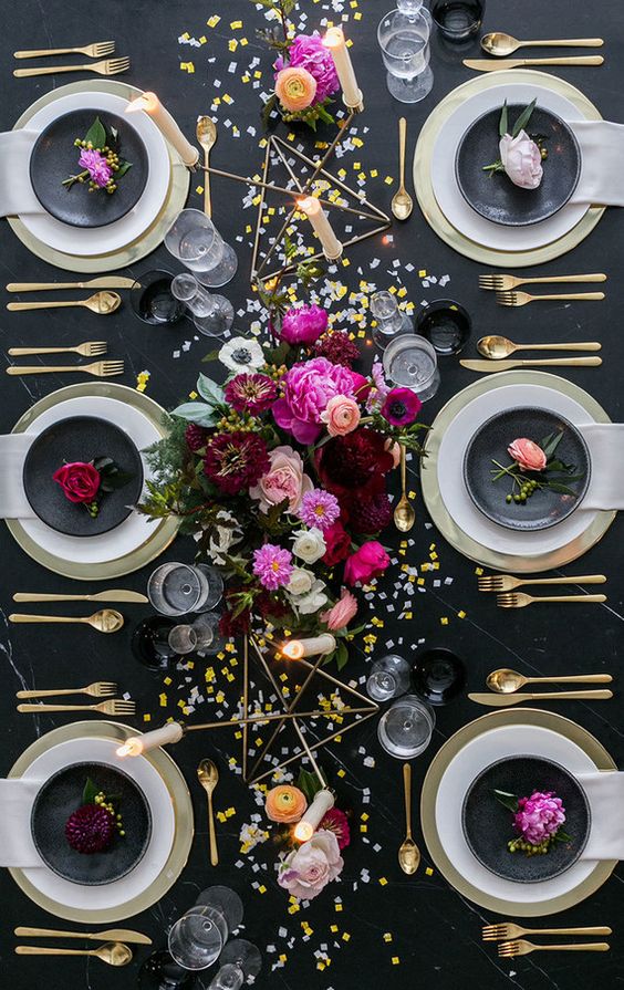 a black and gold NYE table setting with gold chargers and cutlery, gold candleholders, bright blooms and gold and silver sequins on the table