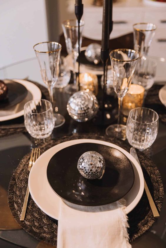 a black and silver NYE tablescape with black printed placemats, black and white plates, black candles and silver disco balls