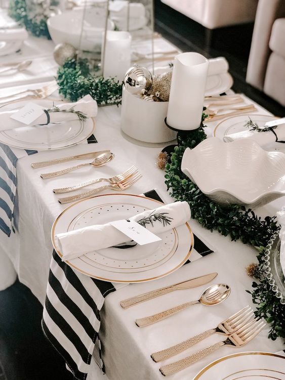 a black and white NYE party table with striped napkins, a greenery runner and pillar candles, silver ornaments and white napkins