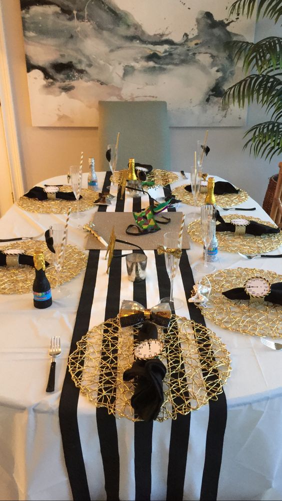 a black, gold and white NYE tablescape with a striped runner, gold placemats, black napkins and clocks, greenery and mini champagne bottles