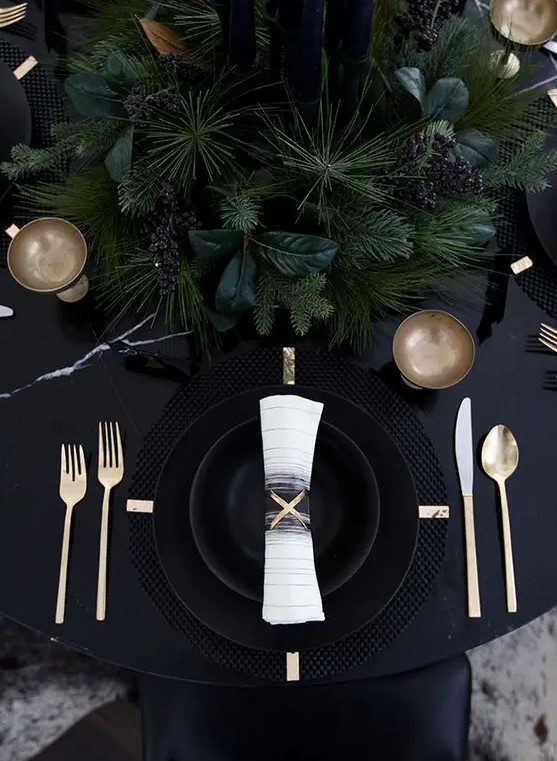 a black table with black plates and cahrgers, lush evergreens and gold cutlery for a moody tablescape
