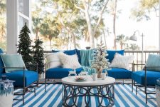 a blue and white coastal Christmas terrace with blue furniture, neutral pillows, Christmas trees, deer and a striped rug