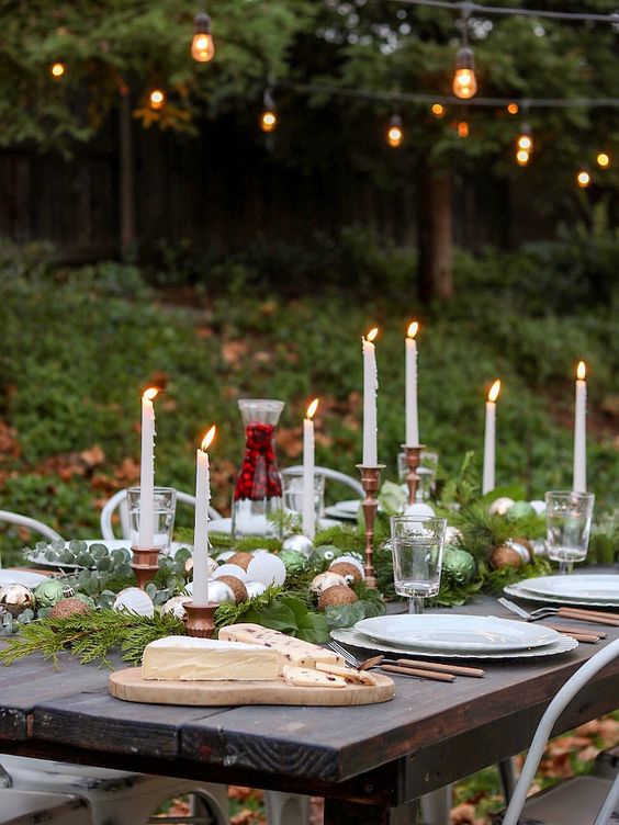 a bold Christmas tablescape with an evergreen runner and ornaments, tall and thin candles in metallic candleholders and neutral porcelain