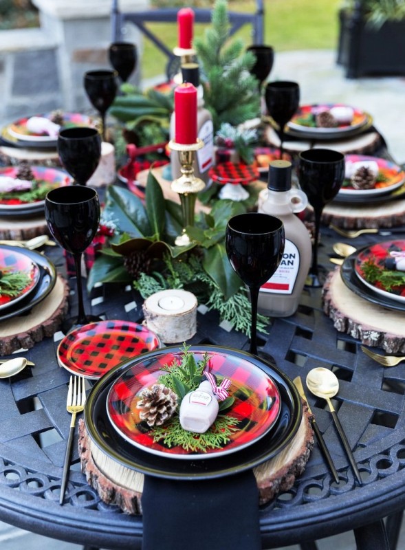 a bold Christmas tablescape with plaid plates and red candles, evergreens and magnolia leaves plus black napkins and glasses