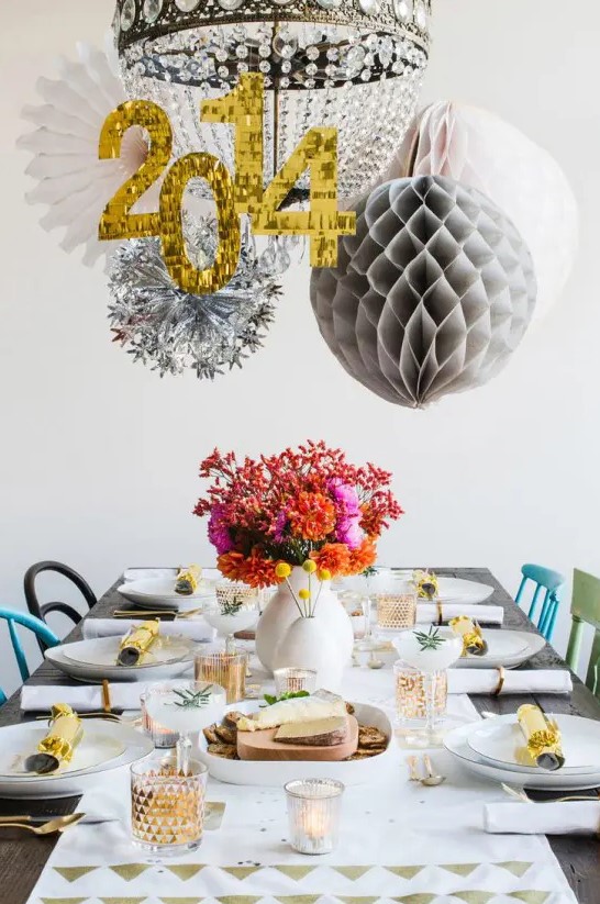 a bright NYE party tablescape with a printed table runner, white porcelain, a bright floral centerpiece, a chandelier decorated with paper pompoms and snowflakes and numbers of the year