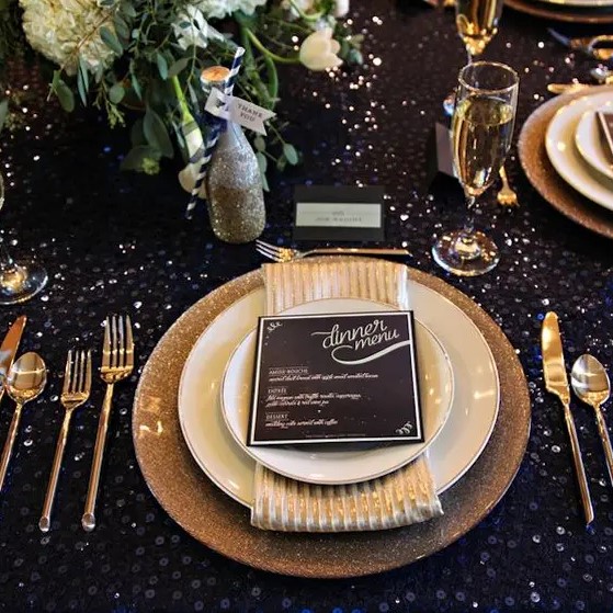 a dark sequin tablecloth, gold glitter chargers, gold cutlery and a lush greenery and bloom centerpiece