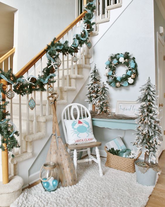 a fantastic beach Christmas nook with an evergreen wreath with ribbons and metallic ornaments and a matching garland on the railing, flocked Christmas trees and a candle lantern with ornaments