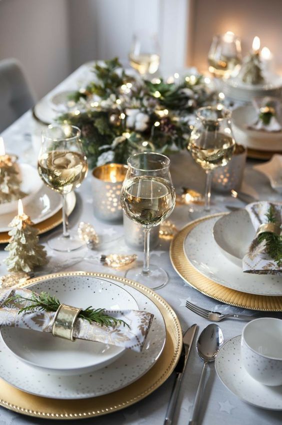 a glam NYE party table in neutrals, with golc chargers and gold candleholders, greenery, gilded Christmas trees and napkin rings