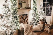a neutral farmhouse living room with a fireplace with firewood, a cluster of flocked Christmas trees and some pillows and blankets