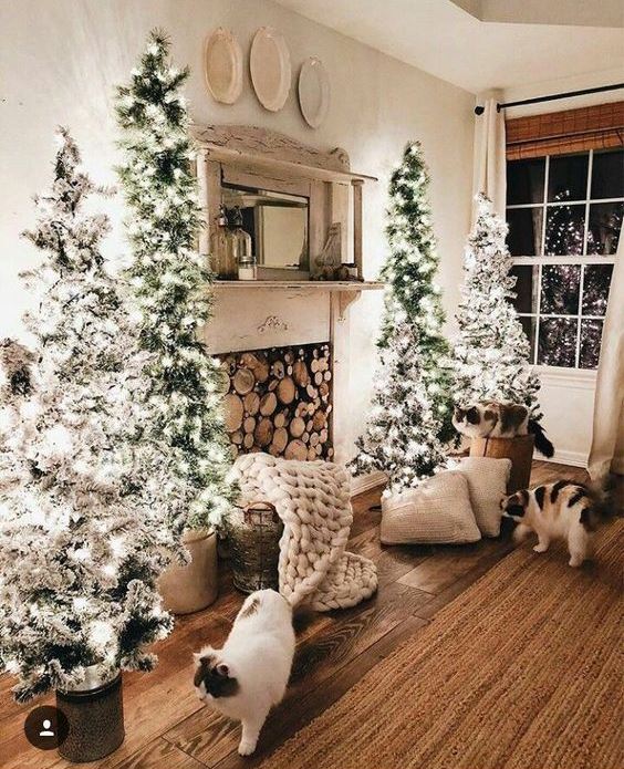 a neutral farmhouse living room with a fireplace with firewood, a cluster of flocked Christmas trees and some pillows and blankets