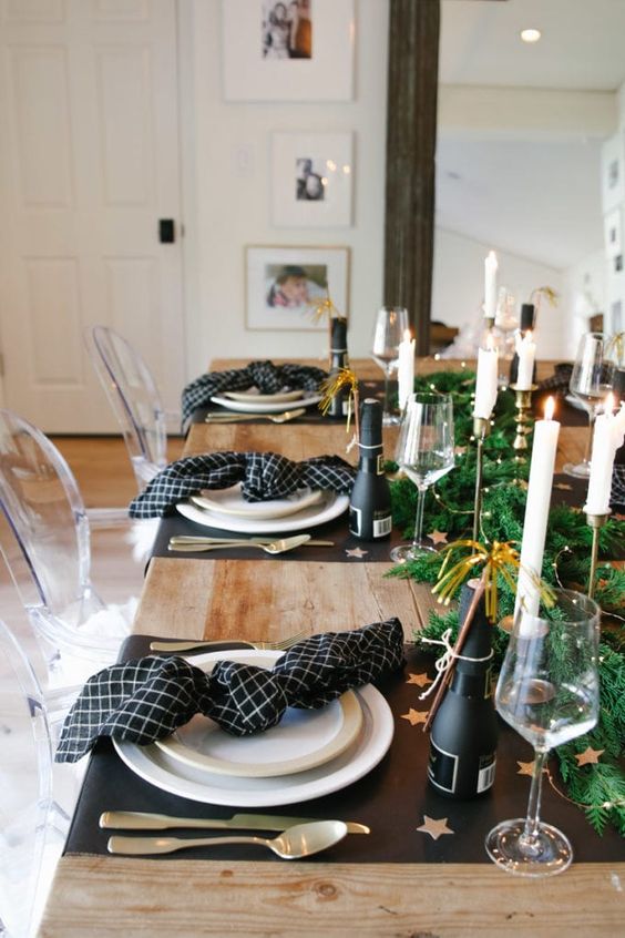 a pretty NYE tablescape with black placemats and printed black napkins, a greenery runner with LEd lights and champagne bottles