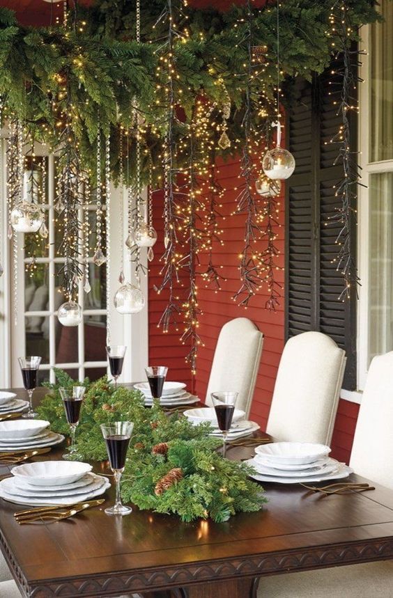 a pretty and elegant outdoor Christmas tablescape with an evergreen and pinecone runner with lights, white porcelain and lights over the table