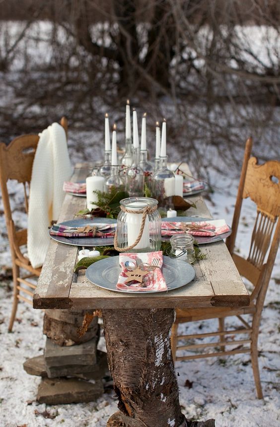 a rustic outdoor Christmas table setting with tall and thin candles, metallic chargers and printed napkins and wooden stars, evergreens on the table