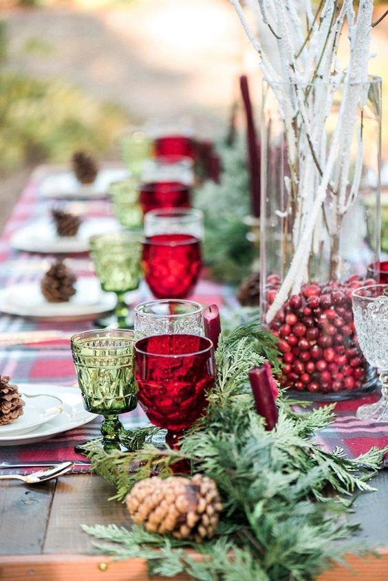 a rustic Christmas tablescape with an evergreen runner, a vase with cranberries and whitewashed branches, red and green glasses