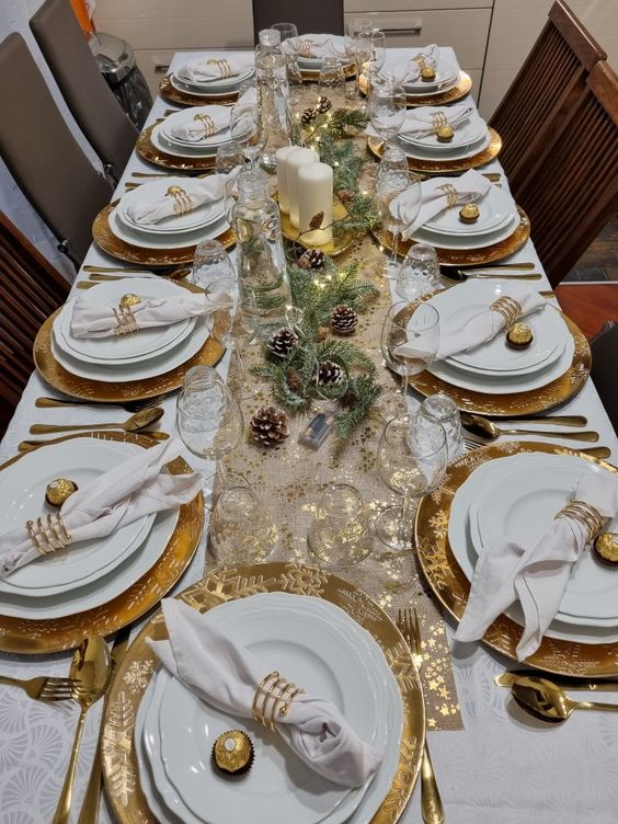 a shiny NYE tablescape with a printed table runner, gold chargers, white linens and gld cutlery plus evergreens and pinecones