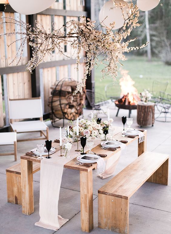 a sophisticated holiday party tablescape with a blush runner, black glasses, a neutral floral centerpiece and tall and thin candles, a blooming overhead installation