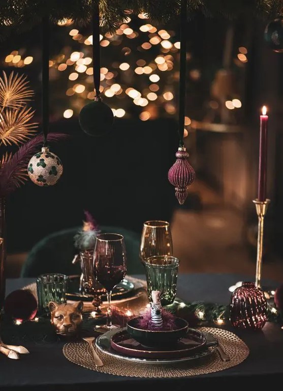 a sophsticated NYE party tablescape in jewel tones, with Christmas ornaments hanging over the table, gold placemats, gold candleholders and purple candles