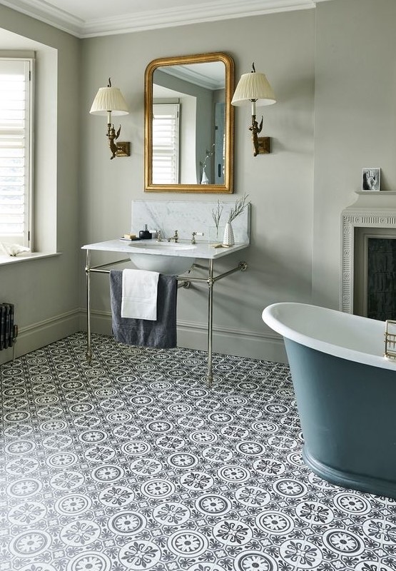 a vintage bathroom with grey walls, a slate blue bathtub, a stand with a sink, a mirror in a gilded frame, sconces and a fireplace plus Moroccan tiles on the floor