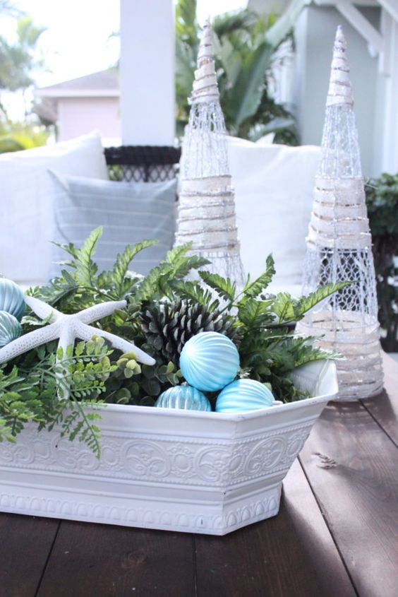 a white bowl with greenery, starfish, pinecones and blue ribbon Christmas ornaments is a lovely Christmas centerpiece for a beach space
