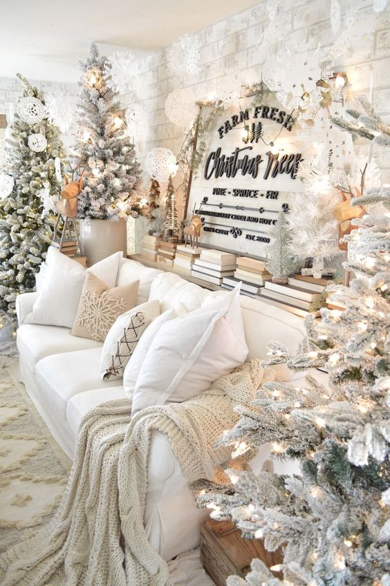 a winter wonderland living room with several flocked Christmas trees with lights, some more mini trees and lots of white paper snowflakes