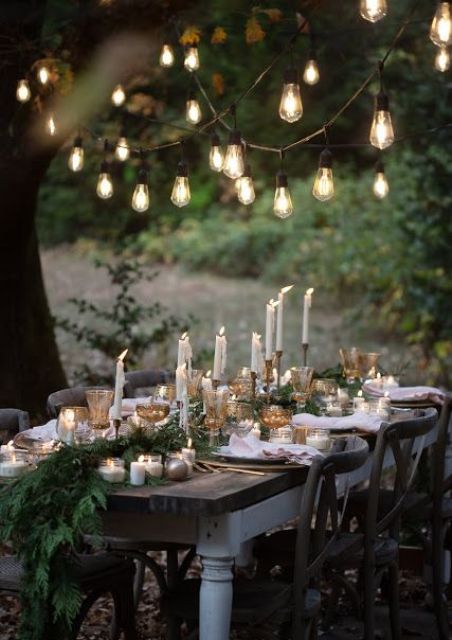 an elegant Christmas tablescape with an evergreen runner, tall and thin candles, metallic ornaments and some lights over the table