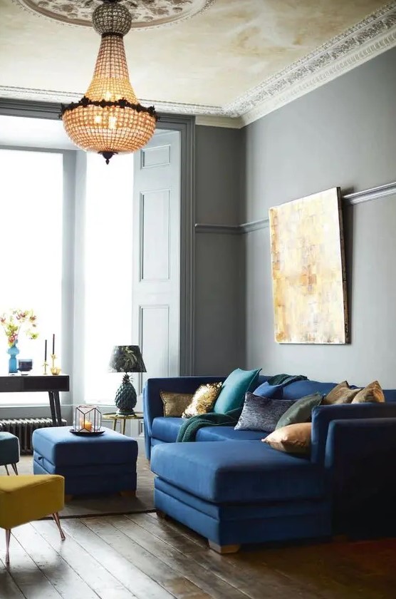 a beautiful and refined living room with grey walls, a navy mid-century modern sectional, a navy pouf and yellow and blue stools, a crystal chandelier and a statement artwork