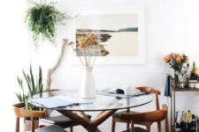 a beautiful mid-century modern dining nook with a round glass table, black chairs, a print, greenery, a bar cart and a cowhide rug