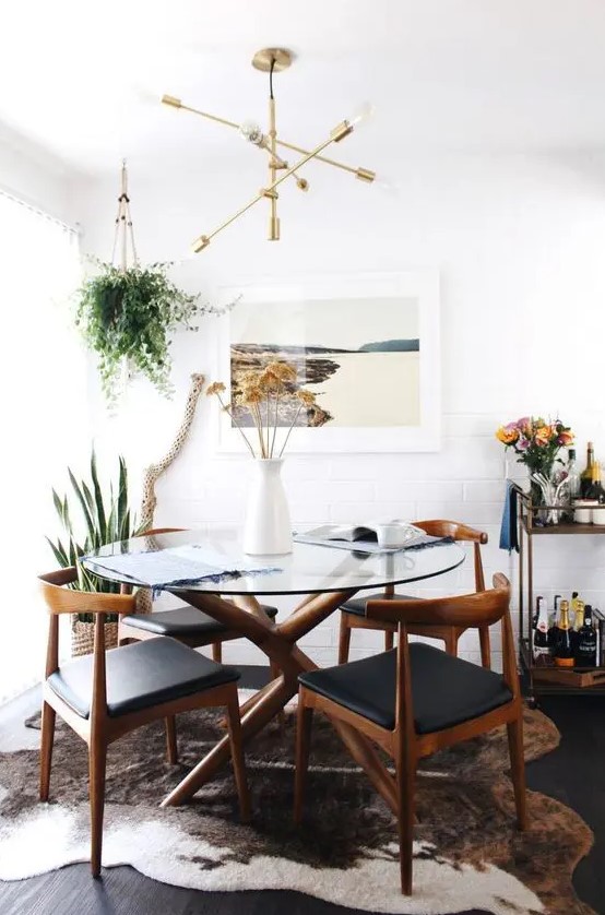 a beautiful mid-century modern dining nook with a round glass table, black chairs, a print, greenery, a bar cart and a cowhide rug