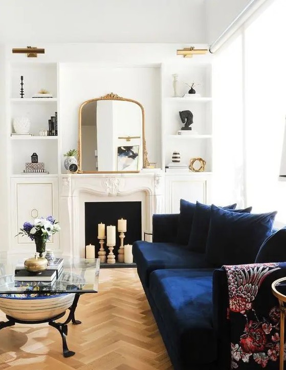 a beautiful neutral living room with a candle fireplace, a modern navy sofa, built-in shelves, a glass coffee table and lovely gold touches here and there