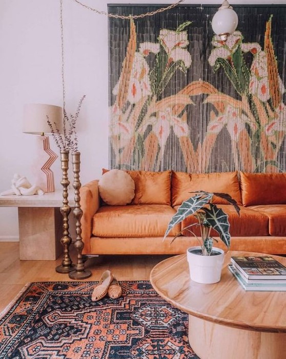 a boho living room with a floral tapestry, an orange sofa, a round table and a side table plus candle holders and a boho rug