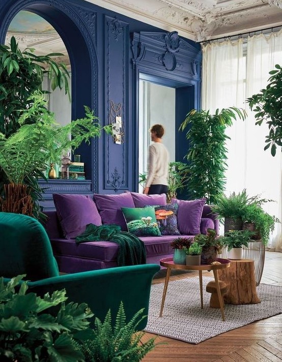 a bright living room with bold blue walls, a purple sofa, an emerald chair, potted greenery and a large mirror