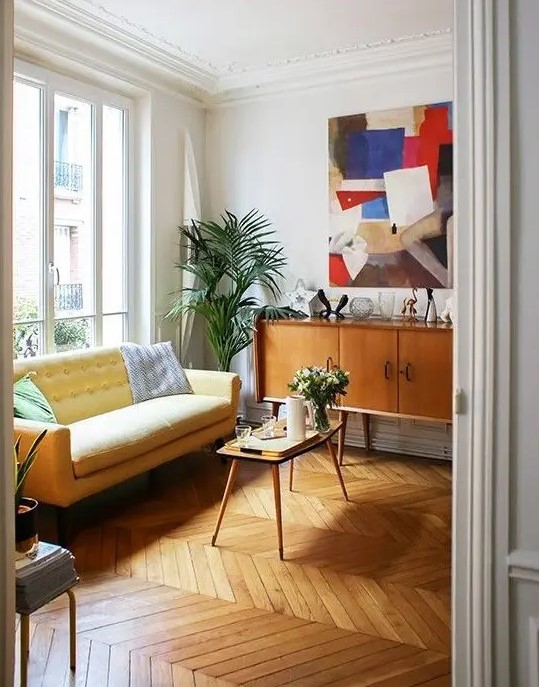 a bright mid-century modern living room with a chevron floor, a yellow loveseat, a stained credenza and a table, a bold artwork