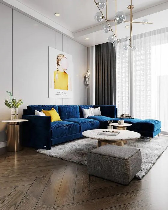 a chic and beautiful living room with dove grey walls, a modern navy sofa, round tables, a grey pouf, a lovely gold chandelier and some touches of yellow