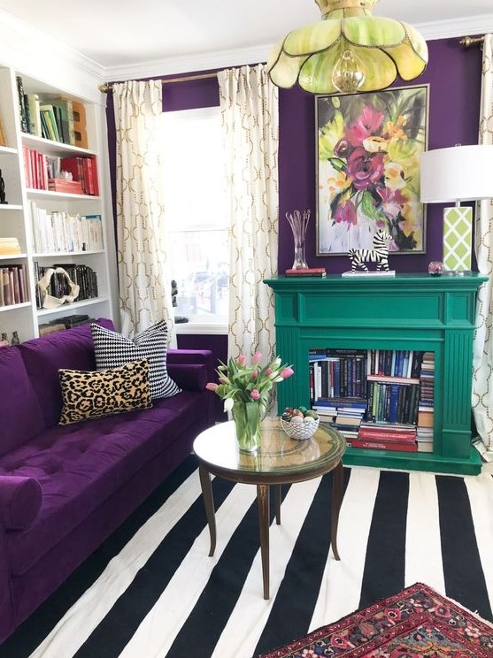 a colorful living room with a depe purple accent wall and a matching sofa, a striped rug, a floral pendant lamp and an emerald fireplace with books