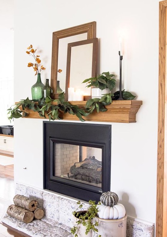 a cool rustic fall mantel with a greenery garland, green bottles with dried blooms, empty frames, mirrors, firewood and stacked heirloom pumpkins